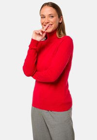 Pull encolère rouge en maille légère, avec des manches longues et un ourlet côtelé. Associé à un pantalon gris, mettant en valeur une coupe ajustée.