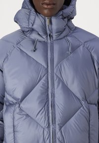 Chaqueta tipo bomber en azul claro, con patrón de costura en diamante, cuello alto, cordones ajustables y cierre frontal con cremallera.