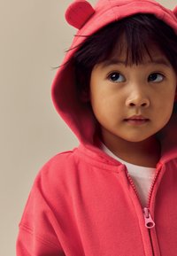 Lyserød fleece-hættetrøje med lynlås og bjørneører, der har en lynlås foran og en blød tekstur, båret over en hvid skjorte, vist mod en neutral baggrund.