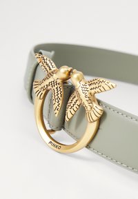 Zaļš ādas josta ar apaļu zelta sprādzi, kurā ir divi rūpīgi izstrādāti putni, kas skatās viens uz otru. Redzama detalizēta šuvuma apdare.