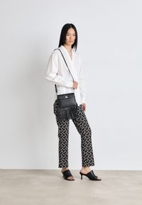 Black leather crossbody bag with fringes, silver hardware, and a snap closure. Worn with a white shirt and patterned black pants.