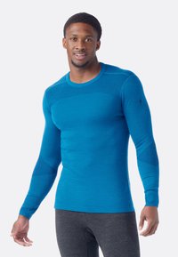Blue long-sleeve thermal shirt with a smooth texture, crew neck, and patterned accents on sleeves, worn over gray pants.