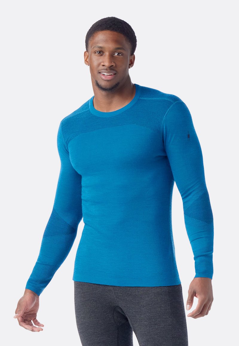 Blue long-sleeve thermal shirt with a smooth texture, crew neck, and patterned accents on sleeves, worn over gray pants.