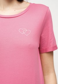 Ροζ πουκάμισο με κοντά μανίκια και δύο μικρές λευκές καρδιές που επικαλύπτονται, ραμμένες στην αριστερή πλευρά του στήθους.