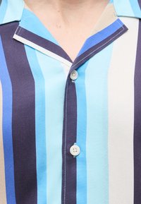 Πουκάμισο με γιακά και κάθετες ρίγες σε ανοιχτό μπλε, σκούρο μπλε, μπεζ και λευκό, με λευκά κουμπιά και εμφανή ραφές.