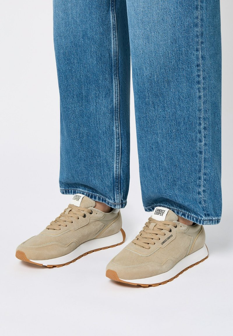 Beige sneakers featuring perforated texture, round toe, flat laces, and a white sole. Paired with loose-fit blue denim jeans.