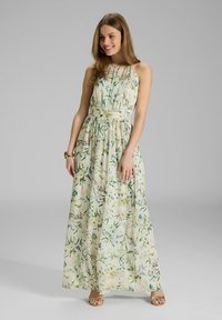 Giovane donna sorridente che indossa un vestito senza maniche, lungo fino al pavimento, di colore crema con stampa floreale multicolore e sandali dorati, in piedi su uno sfondo grigio.