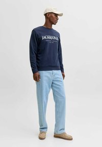 Mørkeblå sweatshirt med hvid logo tekst, kombineret med lyseblå denimjeans og beige sko. Modellen har en lysefarvet kasket på.