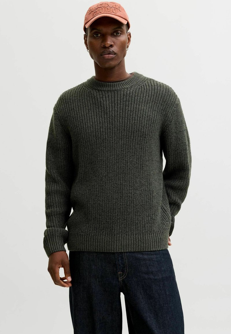 Dark green knitted sweater with a ribbed texture, round neckline, and long sleeves, paired with dark denim jeans and a pink cap.