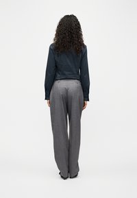 Άτομο με σγουρά μαλλιά που φοράει σκούρο πουκάμισο με μακριά μανίκια και γκρι φαρδιά παντελόνια, όρθιο με την πλάτη στραμμένη σε ένα απλό λευκό φόντο.