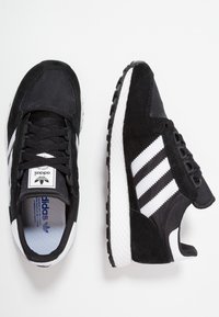 Mustat ja valkoiset Adidas-tennarit, joissa on nauhakiinnitys, kolme raitaa sivuilla ja teksturoitu valkoinen välipohja valkoista taustaa vasten.