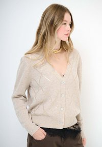 Cardigan beige con scollo a V, caratterizzato da un motivo a diamante in marrone chiaro e fili metallici. Ha chiusure con bottoni e polsini a costine.