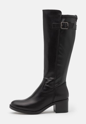 Botte en cuir noire montant jusqu'au genou avec un petit talon carré et une lanière ajustable avec boucle près du haut, sur un fond uni.