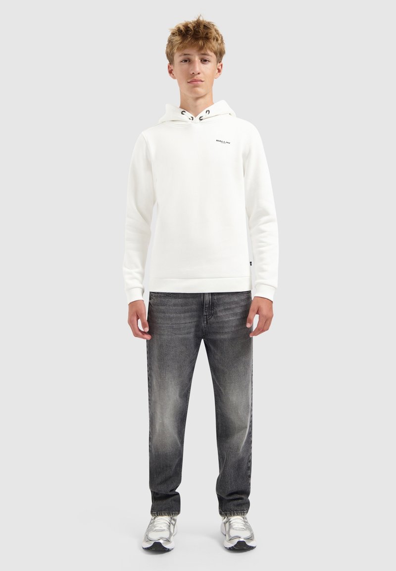 Jonge man staat, gekleed in een witte hoodie met een klein tekstlogo, vervaagde zwarte jeans en witte sneakers, tegen een effen lichte achtergrond.