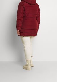 Burgunderfärgad pufferjacka med horisontell söm, huva och en något avsmalnande passform, kombinerad med ljusbeiga byxor och kraftiga beiga stövlar.