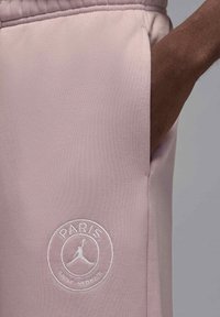 Lähikuva henkilön kädestä vaaleanpunaisten housujen taskussa, joissa on valkoinen Paris Saint-Germain -logo kirjailtuna taskun läheisyyteen.