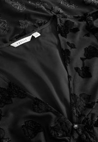 Czarna tkanina z kwiecistym wzorem z koronki, z gładkim wnętrzem i metką z napisem "NOELLA". Widoczny zapięcie na guziki.