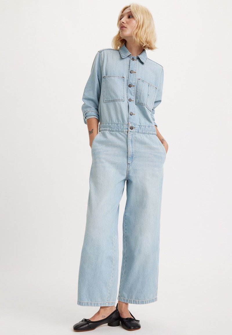 Lyseblå denim-jumpsuit med knapper foran, spidst krave, to brystlommer og viddeben. Stylet med sorte flade sko.