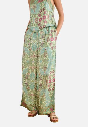 Mujer con pantalones sueltos de talle alto con estampados florales en verde, azul y rosa, y sandalias doradas de tiras, con las manos en los bolsillos.