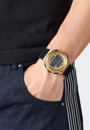 Digitalni sat sa zlatnim kućištem, crnim gumennim remenom i velikim zaslonom koji prikazuje vrijeme. Nositi na zapešću, blizu plavog traper džepa.