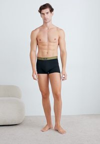 Boxer-briefs noirs avec une ceinture verte, design ajusté et texture lisse. Le modèle est pieds nus sur un sol de couleur claire.