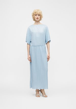 Mujer con cabello rizado y rubio, vistiendo un vestido midi plisado azul claro y sandalias beige de tacón alto, posando frente a un fondo blanco liso.