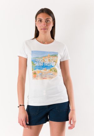 Giovane donna con una maglietta bianca con scena costiera di Saint Tropez e pantaloncini blu scuro, in piedi contro uno sfondo bianco neutro.