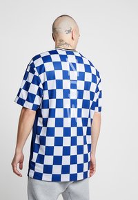 Modrý a bílý kostkovaný dres s krátkým rukávem a síťovanou texturou, na zadní straně se vertikálním designem "N" a s kulatým výstřihem.