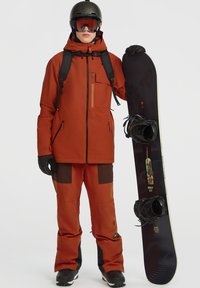 Snowboarder in een oranje jack en broek met zwarte accenten, die een zwart snowboard vasthoudt. Het jack heeft een voorkant rits en een borstzak.