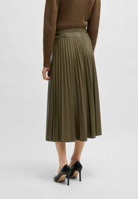 Olivgrüner, plissierter Rock mit hohem Bund, aus einem glatten Material, kombiniert mit schwarzen High Heels.