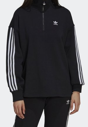 Chándal negro de Adidas con tres franjas blancas en las mangas y los pantalones, con cremallera corta en la parte superior y logotipos de trébol en el pecho y el muslo.