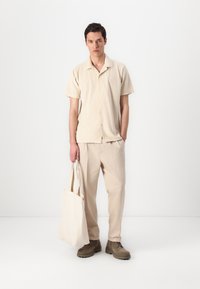 Μπεζ κοντομάνικο πουκάμισο με κουμπιά μπροστά, συνδυασμένο με ανοιχτό μπεζ παντελόνι και τσάντα καμβά. Καφέ μποτάκια με κορδόνια ολοκληρώνουν το σύνολο.