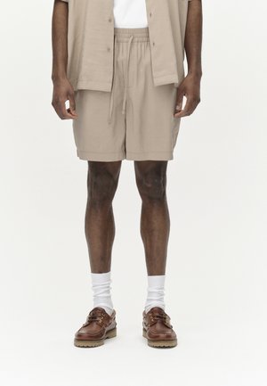 Beige shorts with an elastic waistband and drawstring, paired with white socks and brown loafers. The outfit has a relaxed fit and design.