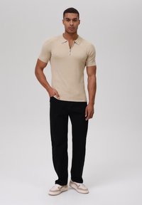 Beige geripptes Poloshirt mit kurzen Ärmeln und Reißverschlusskragen, kombiniert mit schwarzen geraden Hosen und beigen Sneakers mit weißen Akzenten.