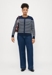 Μπλέ και λευκή ριγέ κάρντιγκαν με κουμπί, συνδυασμένη με μπλε παντελόνια φαρδιάς γραμμής. Διαθέτει ριμπ ύφανση στο τελείωμα και μακριά μανίκια.