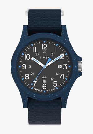 Zegarek Timex z teksturowanym, ciemnoniebieskim paskiem i kopertą z silikonu. Czarna tarcza ma białe i niebieskie znaczniki godzin, białe wskazówki oraz wyświetlacz daty.