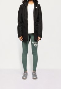 Giacca nera impermeabile con cappuccio, t-shirt bianca, leggings verdi con logo e scarpe sportive grigie; abbigliamento attivo casual.