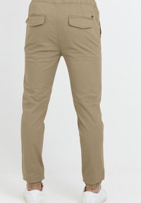 Beżowe spodnie cargo z dwiema tylnymi kieszeniami, zwężanymi nogawkami i elastycznymi mankietami, zestawione z białymi trampkami na jednolitym tle.