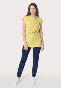 Żółta, zwisająca, krótko rękawowa bluzka z dekoltem cowl, zestawiona z ciemnoniebieskimi skinny jeansami i białymi trampkami. Gładka tekstura, casualowy styl.