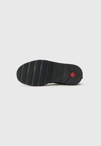 Semelle de chaussure en caoutchouc noir avec une surface texturée, présentant des rainures horizontales et un accent circulaire rouge près du talon.
