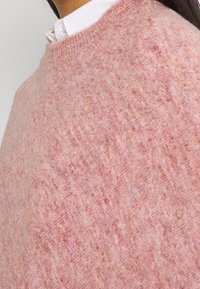 Pull en maille rose présentant une texture douce, un col rond et une coupe légèrement ample. Le tissu montre des variations subtiles de couleur.