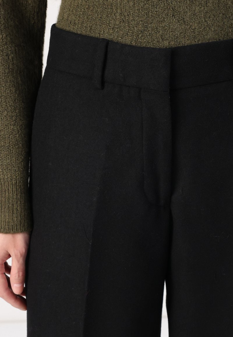 Pantaloni di lana nera con un fit su misura, dotati di una parte frontale piatta, zip nascosta e tasche laterali. Finitura testurizzata e cintura robusta.