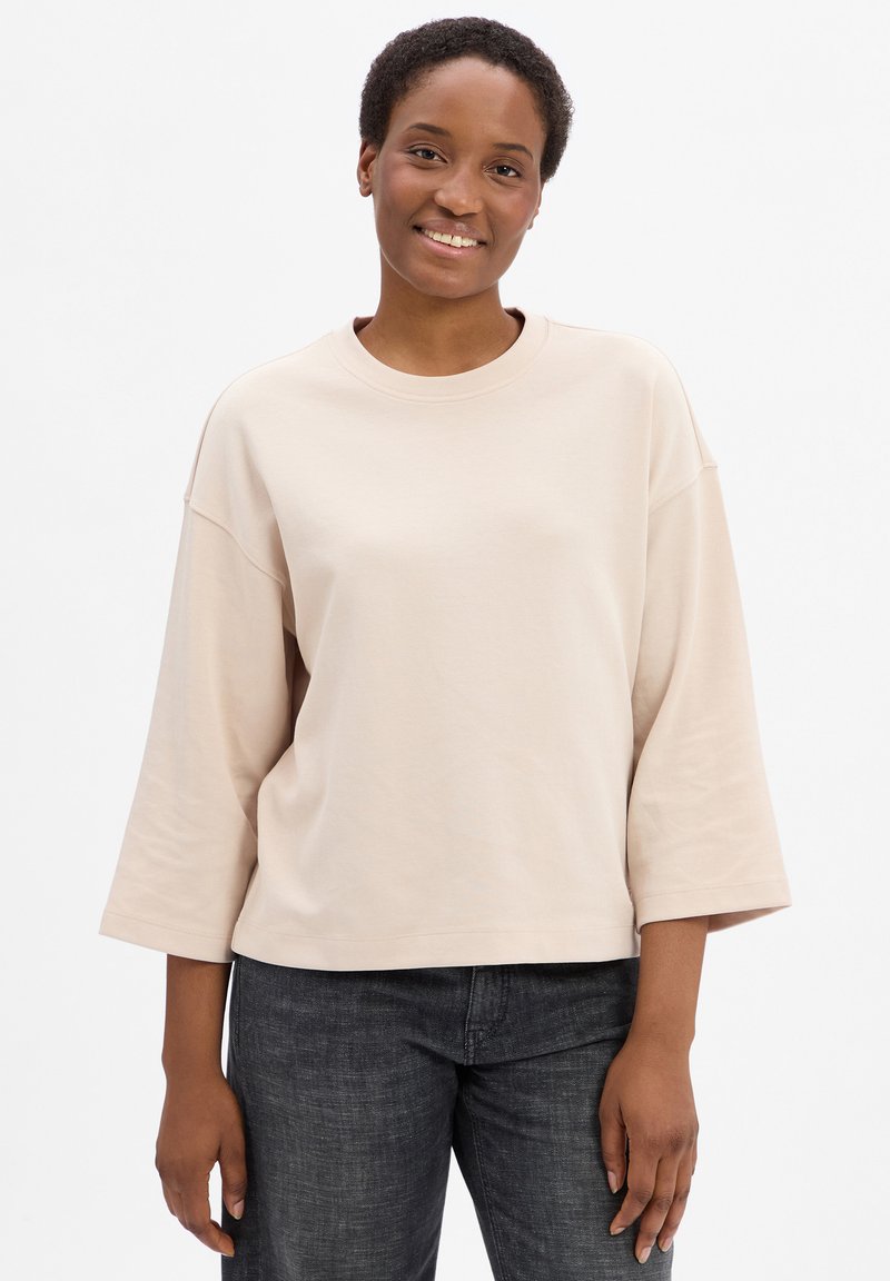 Helles, beiges oversized Sweatshirt mit Rundhalsausschnitt und Dreiviertelärmeln, kombiniert mit dunkelgrauen Jeans. Aus glattem Baumwollmaterial.
