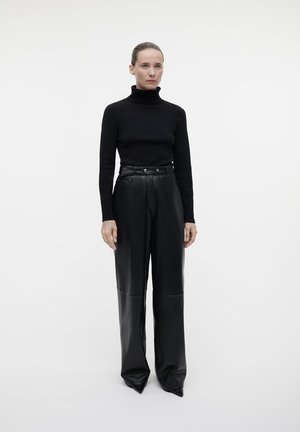 Maglione a collo alto nero abbinato a pantaloni in pelle a vita alta e gamba larga. Texture lisce, design strutturato e scarpe con punta.