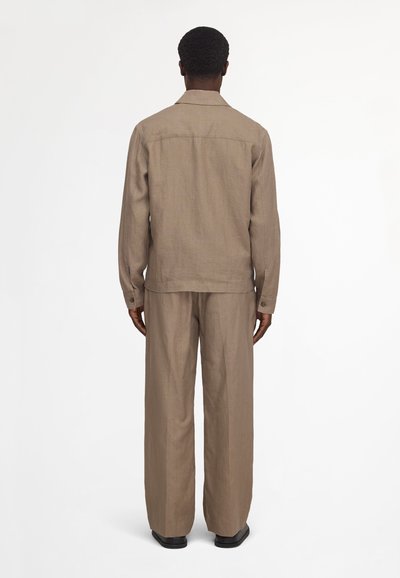 Homme debout, de dos, portant une chemise marron à manches longues et un pantalon ample assorti, avec des chaussures noires, sur un fond blanc uni.