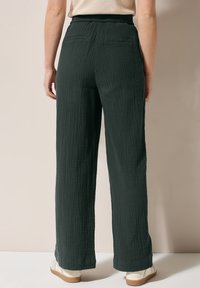 Tmavě zelené texturované kalhoty širokého střihu s elastickým pasem, s kapsami na zadní straně, kombinované se světlými botami na neutrálním pozadí.