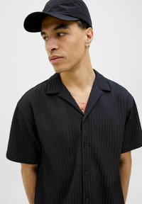 Chemise noire à manches courtes avec un design texturé et côtelé, dotée d'un col ouvert. Comprend des boutons noirs et une coupe décontractée. Portée avec une casquette assortie.