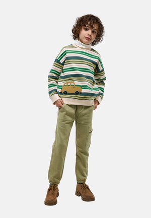 Enfant aux cheveux bouclés portant un pull rayé vert avec un écusson de voiture jaune, un pantalon cargo vert clair et des chaussures marron à lacets, debout les mains dans les poches.