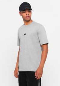 Sivá bavlnená tričko s krátkymi rukávmi a okrúhlym výstrihom; na hrudi je umiestnené malé čierne logo Adidas. Spárované s čiernymi nohavicami.