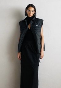 Kvinne som har på seg klare briller, svart ermeløs Nike vest, svart turtleneck med nøkkelhull, og lang svart skjørt, poserer foran en ensfarget lys vegg.